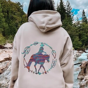 Puede incluir: Una sudadera con capucha beige con un gráfico colorido de una persona montando un caballo en un círculo con plumas y otros elementos decorativos. La sudadera con capucha la lleva puesta una persona de pie frente a un río rocoso.
