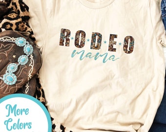May include: A beige t-shirt with the text "RODEO mama" in brown and turquoise letters with a turquoise star pattern. The shirt is on a wooden surface with a pair of brown cowboy boots, a leopard print scarf, and blue jeans.