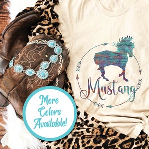 May include: A beige t-shirt with a colorful graphic of a horse in a circle with the word "Mustang" written below. The shirt is on a wooden surface with a pair of brown cowboy boots, a leopard print scarf, and a pair of blue jeans.