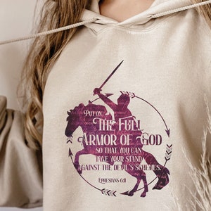 May include: A beige hoodie with a purple graphic print of a knight on horseback. The graphic includes the text "Put on the full armor of God so that you can take your stand against the devil's schemes. Ephesians 6:11"