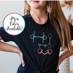 May include: Black t-shirt with a graphic of different horse bits in pink, blue, and teal colors. The text "More Colors Available!" is on a circle in the top left corner.