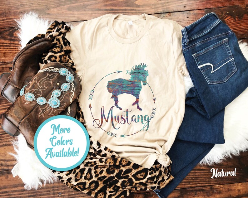 May include: A beige t-shirt with a colorful graphic of a horse in a circle with the word "Mustang" written below. The shirt is on a wooden surface with a pair of brown cowboy boots, a leopard print scarf, and a pair of blue jeans.