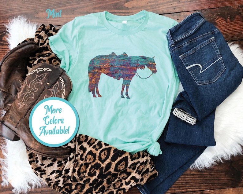 Op de afbeelding: Een turquoise T-shirt met een kleurrijk silhouet van een paard dat een zadel draagt. De tekst "Mint" staat in de linkerbovenhoek van de afbeelding. De tekst "More Colors Available!" staat in een cirkel in de linkerbenedenhoek van de afbeelding.