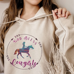 May include: A beige hoodie with a graphic print of a cowgirl riding a horse. The graphic is surrounded by a circle with the text "God Bless Cowgirls" in purple.