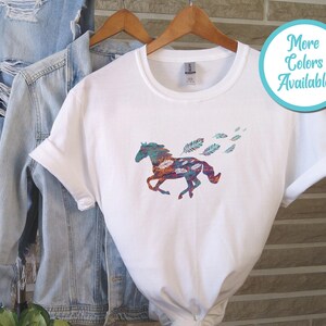 Peut inclure: T-shirt blanc avec un motif de cheval color&eacute; et des plumes. Le cheval court et les plumes sont dispers&eacute;es autour de lui. Le t-shirt est sur un cintre et il y a une veste en jean en arri&egrave;re-plan.