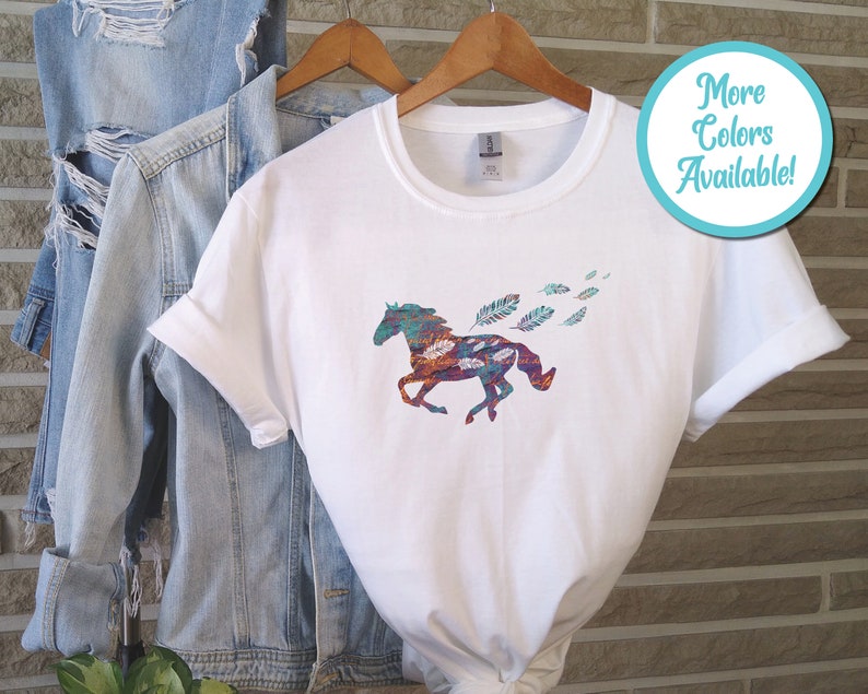 Peut inclure: T-shirt blanc avec un motif de cheval color&eacute; et des plumes. Le cheval court et les plumes sont dispers&eacute;es autour de lui. Le t-shirt est sur un cintre et il y a une veste en jean en arri&egrave;re-plan.