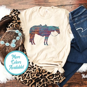 Op de afbeelding: Een beige T-shirt met een kleurrijk silhouet van een paard dat een zadel draagt. De tekst "Meer kleuren beschikbaar!" staat op een blauwe cirkel.