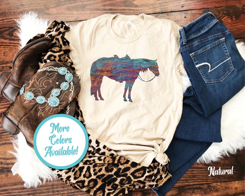 Op de afbeelding: Een beige T-shirt met een kleurrijk silhouet van een paard dat een zadel draagt. De tekst "Meer kleuren beschikbaar!" staat op een blauwe cirkel.
