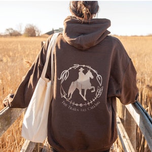 May include: A brown hoodie with a white graphic of a person riding a horse in a circle with the text "The Trails Are Calling".