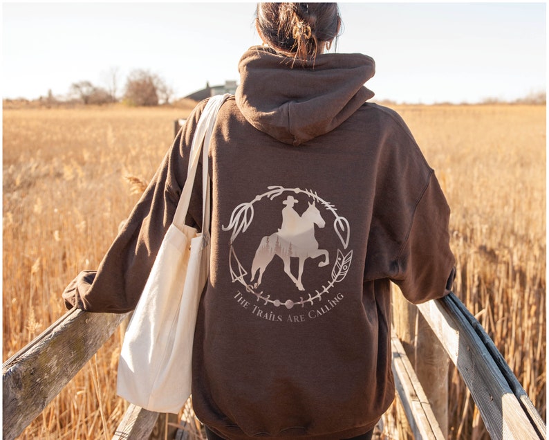 May include: A brown hoodie with a white graphic of a person riding a horse in a circle with the text "The Trails Are Calling".