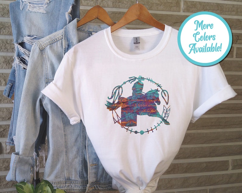 May include: A white short-sleeved t-shirt featuring a graphic of a person riding a horse inside a circular frame. The graphic has a textured, multi-coloured pattern in shades of blue, orange, and purple. A light blue denim jacket hangs behind it.