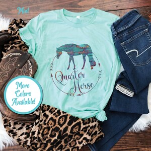 May include: A mint green t-shirt with a silhouette of a quarter horse in a colorful pattern. The shirt has the words "Quarter Horse" in a circle with arrows around it. The shirt is laid out on a wooden surface with a pair of jeans, a cowboy boot, and a leopard print scarf.