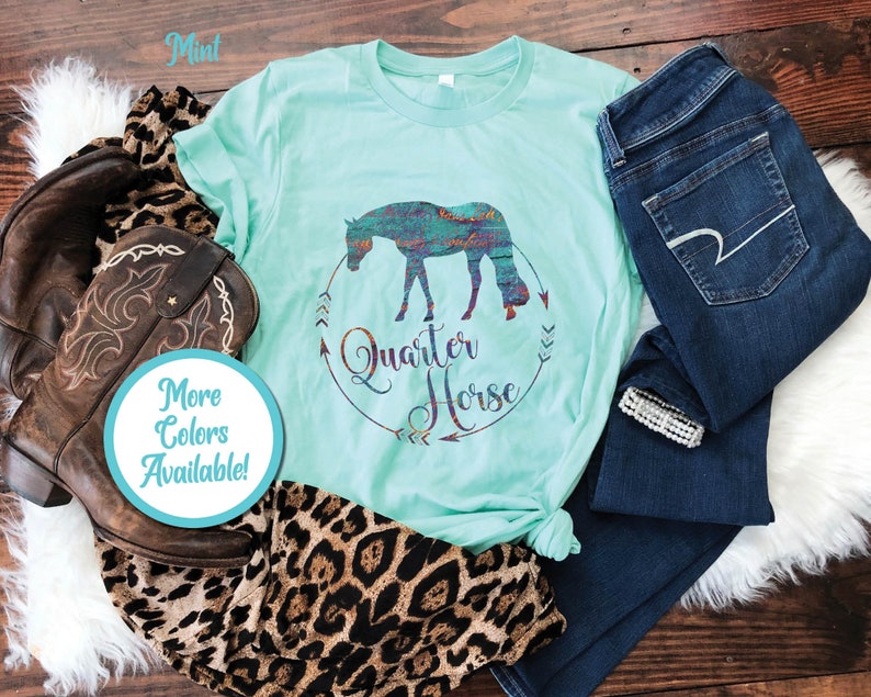 May include: A mint green t-shirt with a silhouette of a quarter horse in a colorful pattern. The shirt has the words "Quarter Horse" in a circle with arrows around it. The shirt is laid out on a wooden surface with a pair of jeans, a cowboy boot, and a leopard print scarf.