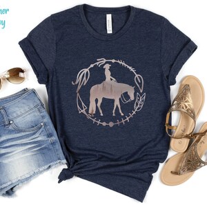 Pode incluir: Uma camiseta azul marinho heather com um gr&aacute;fico de uma mulher montando um cavalo em um c&iacute;rculo com um design floral. O gr&aacute;fico &eacute; de cor ouro rosa.