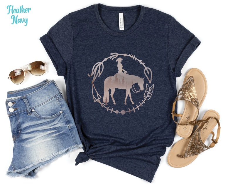 Pode incluir: Uma camiseta azul marinho heather com um gr&aacute;fico de uma mulher montando um cavalo em um c&iacute;rculo com um design floral. O gr&aacute;fico &eacute; de cor ouro rosa.