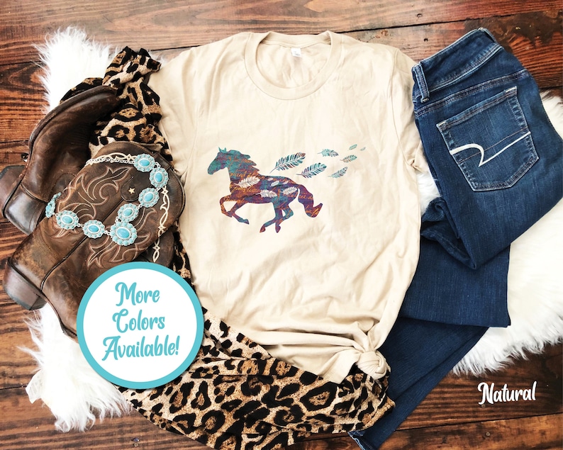 Peut inclure: Un t-shirt beige avec un motif de cheval color&eacute; et de plumes. Le texte "Plus de couleurs disponibles !" est sur un cercle bleu.