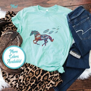 Peut inclure: Un t-shirt vert menthe avec une silhouette de cheval color&eacute;e et des plumes. Le t-shirt est pos&eacute; sur une surface en bois avec un jean, une botte de cowboy et une &eacute;charpe &agrave; imprim&eacute; l&eacute;opard. Le texte "Plus de couleurs disponibles !" est sur un cercle bleu.