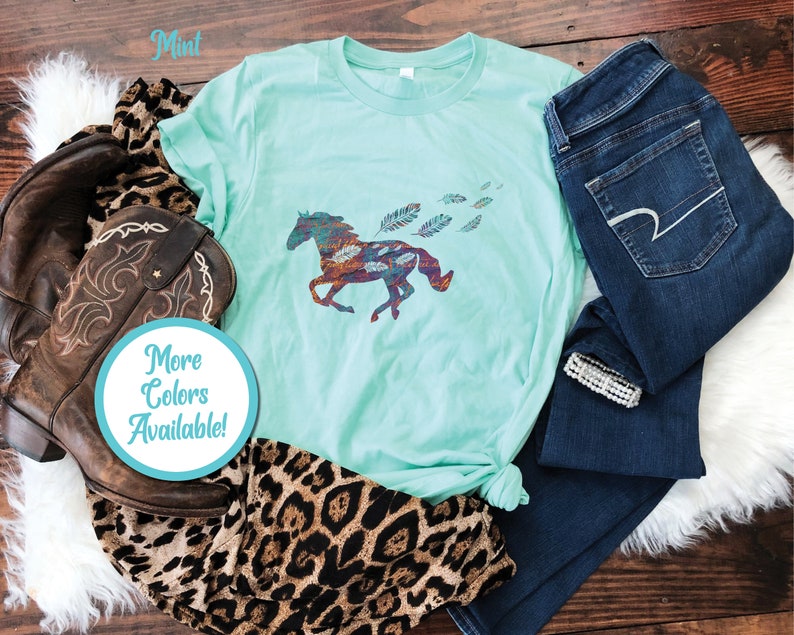 Peut inclure: Un t-shirt vert menthe avec une silhouette de cheval color&eacute;e et des plumes. Le t-shirt est pos&eacute; sur une surface en bois avec un jean, une botte de cowboy et une &eacute;charpe &agrave; imprim&eacute; l&eacute;opard. Le texte "Plus de couleurs disponibles !" est sur un cercle bleu.