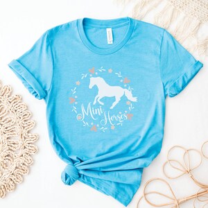 Może przedstawiać: Jasno błękitny t-shirt z białą sylwetką konia i tekstem "Mini Horses" w wianku kwiatowym.