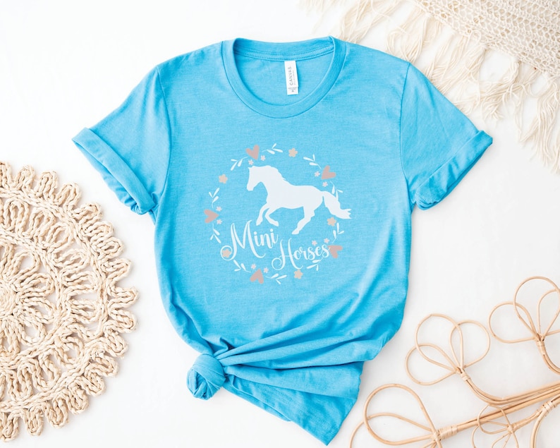 Może przedstawiać: Jasno błękitny t-shirt z białą sylwetką konia i tekstem "Mini Horses" w wianku kwiatowym.