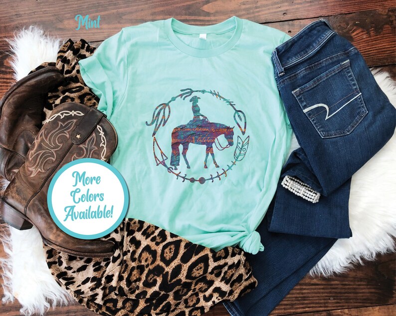 Pode incluir: Uma camiseta verde menta com um gr&aacute;fico de uma mulher montando um cavalo em um c&iacute;rculo com penas e outros elementos decorativos. O texto "Mais cores dispon&iacute;veis!" est&aacute; em um c&iacute;rculo azul no canto inferior esquerdo da imagem.