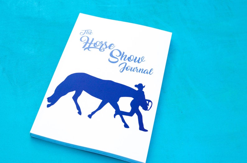 May include: A white journal with a blue silhouette of a horse and rider. The text "The Horse Show Journal" is printed on the cover.
