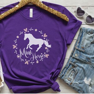 Może przedstawiać: Fioletowy T-shirt z białą sylwetką konia i tekstem "Mini Horses" w r&oacute;żowym wianku kwiatowym.