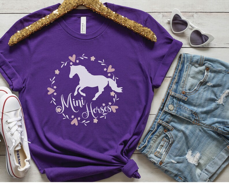Może przedstawiać: Fioletowy T-shirt z białą sylwetką konia i tekstem "Mini Horses" w r&oacute;żowym wianku kwiatowym.