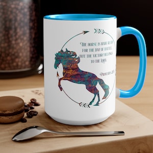 May include: White ceramic mug with a blue rim and handle. The mug features a silhouette of a horse with a textured, colorful design. The text "THE HORSE IS MADE READY FOR THE DAY OF BATTLE BUT THE VICTORY BELONGS TO THE LORD" is printed on the mug, along with the verse "Proverbs 23".