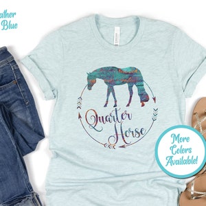 May include: A heather ice blue t-shirt with a graphic of a colorful quarter horse silhouette in a circle with the text "Quarter Horse" and arrows. The text "More Colors Available!" is in a circle below the graphic.