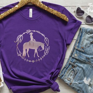 Pode incluir: Camiseta roxa com uma silhueta rosa de uma mulher montando um cavalo em um c&iacute;rculo com flechas e penas. O texto "Team Purple" est&aacute; no canto superior direito da imagem.