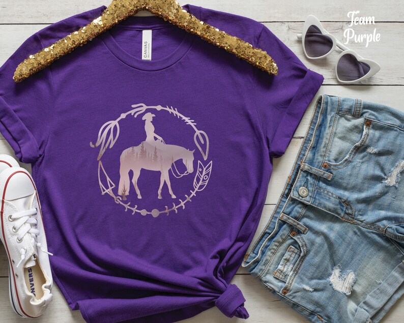 Pode incluir: Camiseta roxa com uma silhueta rosa de uma mulher montando um cavalo em um c&iacute;rculo com flechas e penas. O texto "Team Purple" est&aacute; no canto superior direito da imagem.