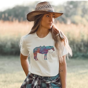Op de afbeelding: Een wit T-shirt met een kleurrijk abstract paardenontwerp. Het paard is zwart omlijnd en heeft een zadel en een hoofdstel. De vrouw draagt een bruine strohoed en een geruit overhemd dat om haar taille is geknoopt.