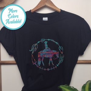 Pode incluir: Camiseta preta com um gr&aacute;fico colorido de uma pessoa montando um cavalo em um c&iacute;rculo com flechas e penas. O texto "Mais cores dispon&iacute;veis!" est&aacute; em um c&iacute;rculo azul no canto superior esquerdo.