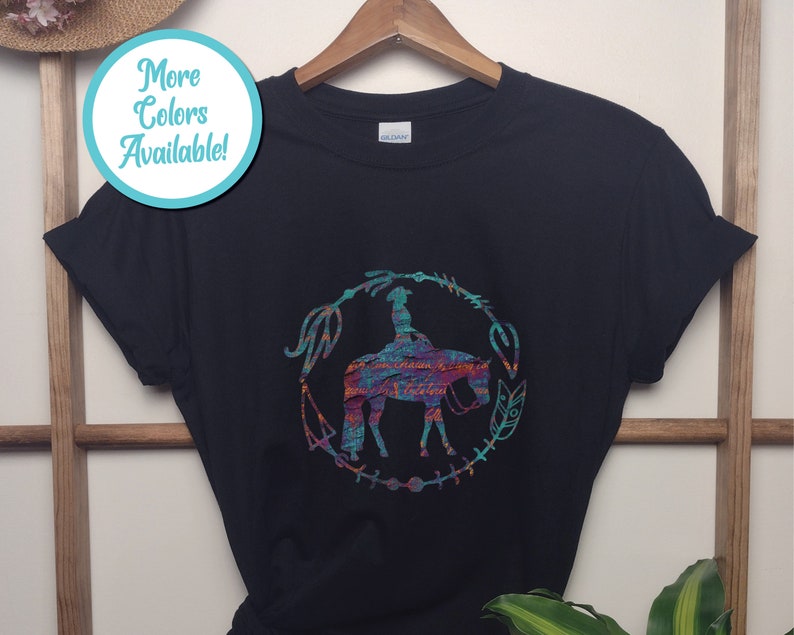 Pode incluir: Camiseta preta com um gr&aacute;fico colorido de uma pessoa montando um cavalo em um c&iacute;rculo com flechas e penas. O texto "Mais cores dispon&iacute;veis!" est&aacute; em um c&iacute;rculo azul no canto superior esquerdo.