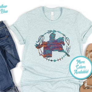 May include: A heather ice blue short-sleeve t-shirt featuring a central graphic of a rider on horseback enclosed within a circular lasso and feather design. The graphic has a textured, multi-coloured appearance with shades of teal, orange, and purple.