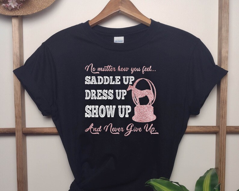 May include: Black t-shirt with a pink glitter design that says "No matter how you feel... Saddle Up Dress Up Show Up And Never Give Up."