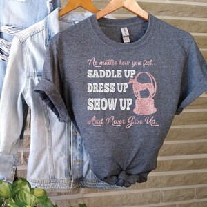 May include: A heather gray t-shirt with a pink glitter design that says "No matter how you feel... Saddle Up, Dress Up, Show Up, And Never Give Up."