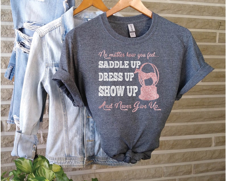 May include: A heather gray t-shirt with a pink glitter design that says "No matter how you feel... Saddle Up, Dress Up, Show Up, And Never Give Up."