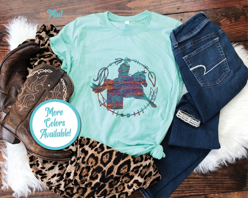 May include: A mint green t-shirt featuring a graphic of a person riding a horse inside a circular lasso frame. The graphic has a textured, multi-coloured appearance with script text. The shirt is displayed with brown leather cowboy boots and dark blue jeans.