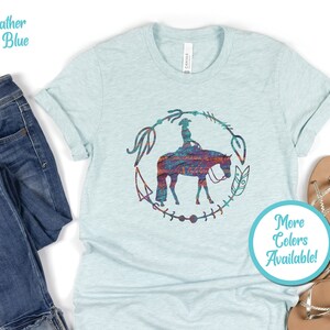Pode incluir: Uma camiseta azul claro com uma silhueta de uma pessoa montando um cavalo em um c&iacute;rculo de flechas e penas. O cavalo e o cavaleiro s&atilde;o um padr&atilde;o colorido de azul, vermelho, laranja e roxo. O texto "Mais cores dispon&iacute;veis!" est&aacute; em um c&iacute;rculo turquesa.