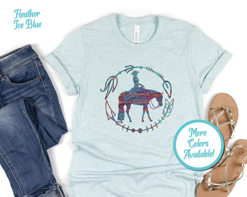 Pode incluir: Uma camiseta azul claro com uma silhueta de uma pessoa montando um cavalo em um c&iacute;rculo de flechas e penas. O cavalo e o cavaleiro s&atilde;o um padr&atilde;o colorido de azul, vermelho, laranja e roxo. O texto "Mais cores dispon&iacute;veis!" est&aacute; em um c&iacute;rculo turquesa.
