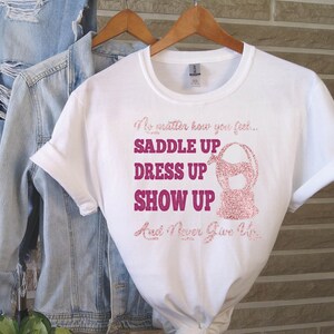 May include: White t-shirt with a pink glitter design that says "No matter how you feel... Saddle Up Dress Up Show Up And Never Give Up".