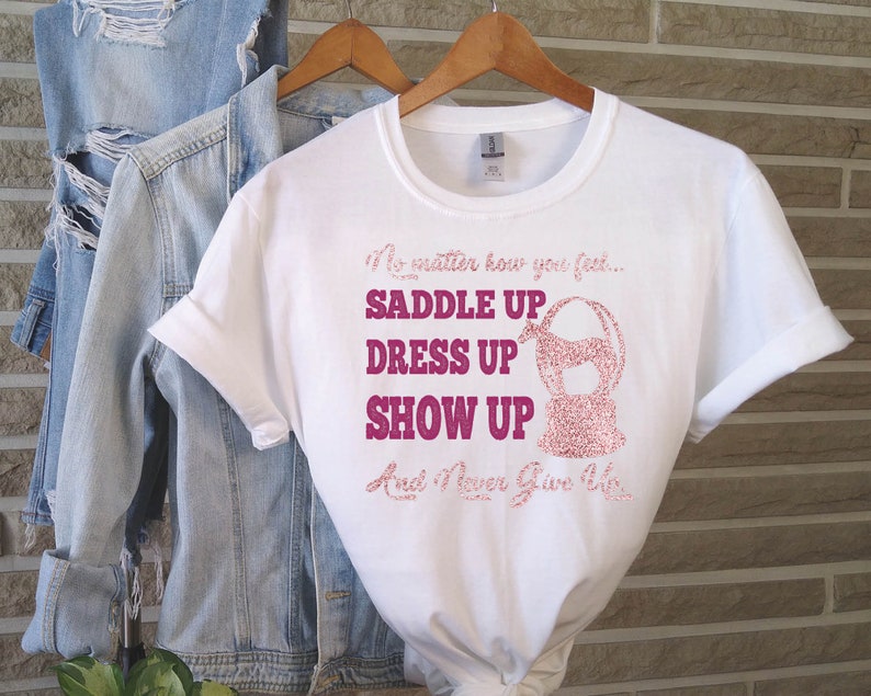 May include: White t-shirt with a pink glitter design that says "No matter how you feel... Saddle Up Dress Up Show Up And Never Give Up".