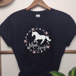 Może przedstawiać: Czarny T-shirt z białą sylwetką konia w r&oacute;żowym wianku kwiatowym. Tekst "Mini Horses" jest napisany r&oacute;żowym pismem.