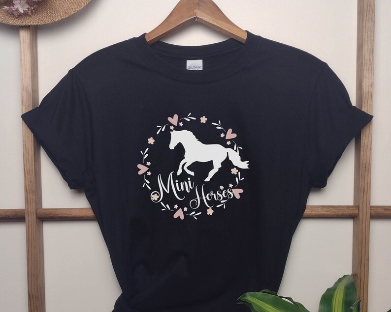 Może przedstawiać: Czarny T-shirt z białą sylwetką konia w r&oacute;żowym wianku kwiatowym. Tekst "Mini Horses" jest napisany r&oacute;żowym pismem.