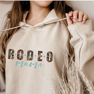 May include: A beige hoodie with a brown and blue patterned design that says "RODEO mama" on the front.