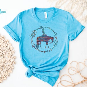 Pode incluir: Uma camiseta azul turquesa com um gr&aacute;fico de uma mulher montando um cavalo em um c&iacute;rculo de flechas e penas. O gr&aacute;fico est&aacute; em um arco-&iacute;ris de cores.