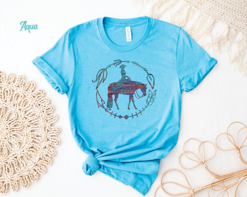 Pode incluir: Uma camiseta azul turquesa com um gr&aacute;fico de uma mulher montando um cavalo em um c&iacute;rculo de flechas e penas. O gr&aacute;fico est&aacute; em um arco-&iacute;ris de cores.