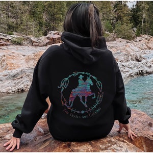 May include: Black hooded sweatshirt with a colorful graphic of a person riding a horse. The graphic is surrounded by a circle of leaves and the text "The Trails Are Calling".